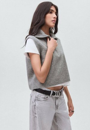 Jonge vrouw in een mouwloze grijze wollen vest over een wit T-shirt en lichtgrijze jeans, kijkend naar de zijkant met haar hand bij de kraag.