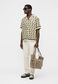 Muž s okuliarmi na slnko, vzorovaná košeľa s krátkym rukávom, biele nohavice, hnedé a béžové topánky, drží veľkú béžovú semišovú tašku na rameno.