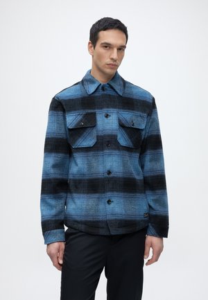 Homme portant une veste à carreaux bleu et noir boutonnée avec des poches à rabat, debout devant un fond clair uni.