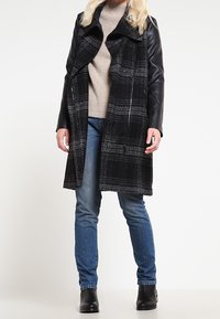 Schwarz-grauer karierter Mantel mit Lederärmeln, offenes Design, kombiniert mit einem beigen Pullover und blauen Jeans, getragen mit schwarzen Stiefeletten.