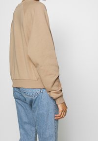 Sweat-shirt beige avec un col rond, des poignets et un ourlet côtelés. Texture douce ; porté avec un jean denim bleu clair avec des poches arrière.