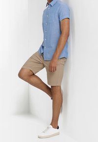 Mężczyzna w jasnoniebieskiej koszuli z krótkim rękawem na guziki, beżowych spodniach krótkich i białych tenisówkach, opierający się o białą ścianę z jedną ugiętą nogą w kolanie.