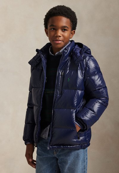 Garçon portant une doudoune bleu marine brillante avec capuche, chemise rayée, pull sombre et jean bleu, debout avec une main dans la poche.