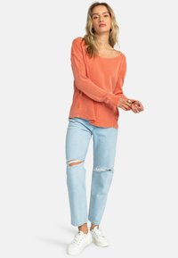 Pehmeästä kankaasta valmistettu korallinen pitkähihainen paita, jossa on pyöreä pääntie, yhdistetty vaaleansinisillä revityillä farkuilla ja valkoisilla tennareilla. Rentoa, löysää istuvuutta.