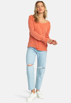Koralowa bluzka z długim rękawem z miękkiego materiału z dekoltem w łódkę, zestawiona z jasnoniebieskimi podartymi jeansami i białymi trampkami. Casualowy, luźny krój.