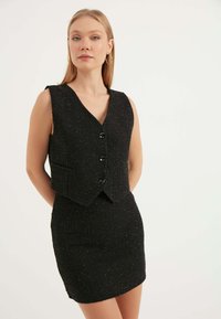 Svart tweedväst med V-ringning, utan ärmar, med tre knappar och en ficka; kombinerad med en figurnära svart minikjol.