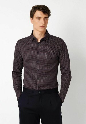 Mann mit einem dunkel gemusterten Hemd mit Knöpfen und schwarzen Hosen steht mit den Händen in den Taschen und schaut leicht zur Seite.