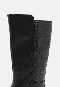 Des bottes noires montantes en cuir lisse et matériau synthétique texturé avec un motif en forme de cœur. Fermeture éclair sur le côté pour un enfilage facile.