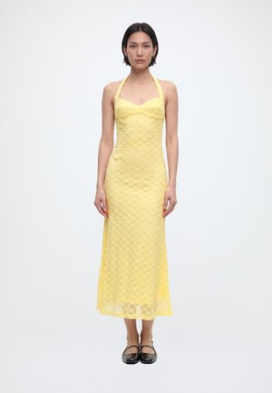 Kobieta stojąca przodem, ubrana w żółtą, teksturowaną sukienkę midi na szyję i czarne buty typu Mary Jane na białym tle.