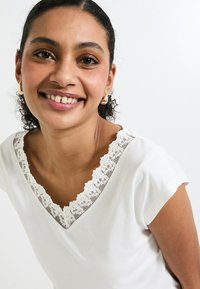 Chemise blanche à col en V avec une finition en dentelle au niveau du col, manches courtes, et tissu doux avec une texture lisse et un léger éclat.