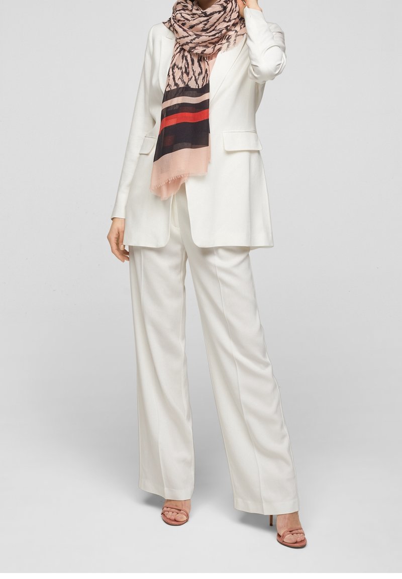 Femme portant un costume blanc ajusté avec un pantalon à jambes larges, une écharpe rose et noire à motifs, et des sandales roses à talons.