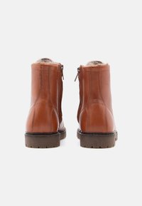 Une paire de bottines en cuir marron avec doublure en fourrure, semelles robustes, coutures visibles et zips latéraux, présentée de l'arrière sur un fond blanc.
