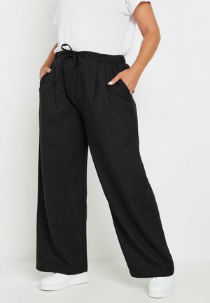 Pantalons larges noirs avec plis, taille élastique et poches latérales. Assortis à des baskets blanches. Texture de tissu lisse et léger.