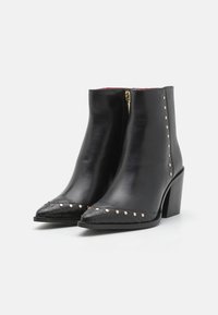 Botas de tobillo de cuero negro con punta afilada, que presentan un detalle texturizado negro y tachuelas doradas a lo largo del borde, y un tacón cuadrado.