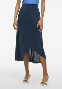 Granatowa spódnica midi z miękkiego, elastycznego materiału, z zamkiem bocznym i rozcięciami, zestawiona z białymi sandałami na otwartym palcu.