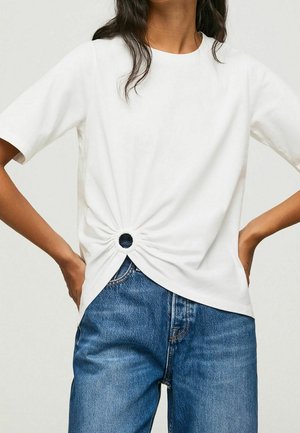 Biała koszulka z krótkim rękawem z ozdobnym skrętem i okrągłym wycięciem w talii, zestawiona z niebieskimi jeansami.