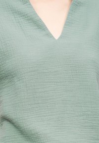 Ανοιχτό πράσινο υφασμάτινο τοπ με υφή και λαιμόκοψη σε σχήμα V, που αφήνει εκτεθειμένη την άνω περιοχή του στήθους και των ώμων.