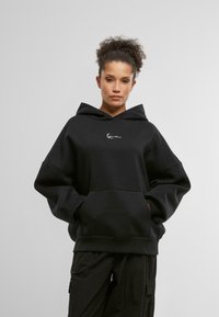Zwarte oversized hoodie van zacht materiaal. Kenmerken zijn een voorzak, een capuchon met aantrekkoord en een wit geborduurd logo op de borst.