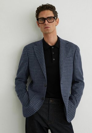 Hombre con cabello corto y rizado y gafas negras, vistiendo un blazer azul con estampado, camiseta polo negra y pantalones negros, de pie contra una pared blanca.