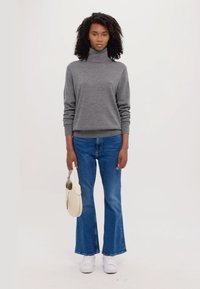 Grå høyhalset genser med lange ermer, kombinert med blå utsvingte jeans. Modellen holder en kremfarget håndveske og har på seg hvite joggesko.