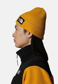Yellow ribbed beanie with a black label featuring a white logo, worn by a person in a black and yellow fleece jacket, shown in profile.
