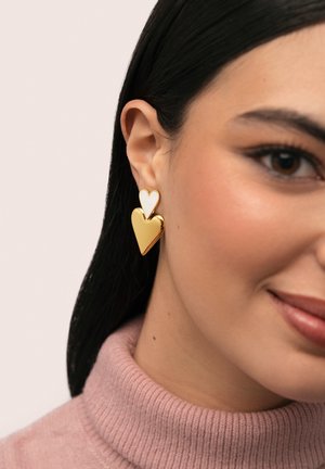 Žena s tmavými vlasy, která nosí zlaté a bílé náušnice ve tvaru dvou srdcí a růžový rolák.