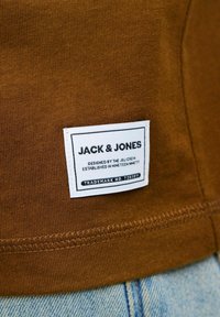Tejido acanalado marrón con una etiqueta blanca que muestra "JACK & JONES" y detalles de diseño. Costuras visibles en el dobladillo y los bordes de la etiqueta.