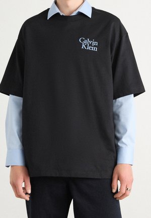 Person trägt ein schwarzes Calvin Klein T-Shirt über einem hellblauen Langarmshirt, schwarze Hose und einen silbernen Ring an der rechten Hand.