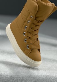 Sneaker alta marrone e morbida con texture soffice, occhielli rotondi e lacci abbinati. Suola in gomma bianca con dettaglio di grip texturizzato.