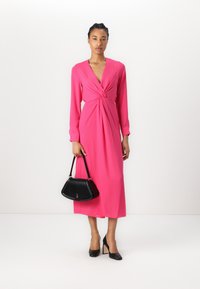 Fuchsia long-sleeve dress with a knotted front detail, paired with a black structured handbag and black block-heeled shoes.