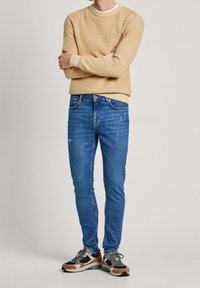 Beige mikina s kulatým výstřihem, modré slim-fit džíny a vícebarevné tenisky s černými akcenty. Měkká látka, neformální design.