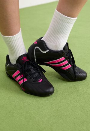 Osoba nosząca białe skarpetki i czarne buty sportowe Adidas z różowymi paskami, stojąca na jasnozielonej podłodze.