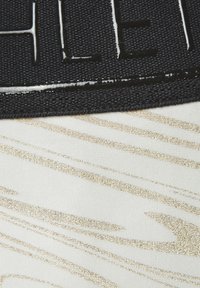 Schwarzes Stoffetikett mit erhabener Schrift, angebracht auf einem strukturierten Hintergrund mit goldenen diagonalen Streifen auf einer weißen Fläche.