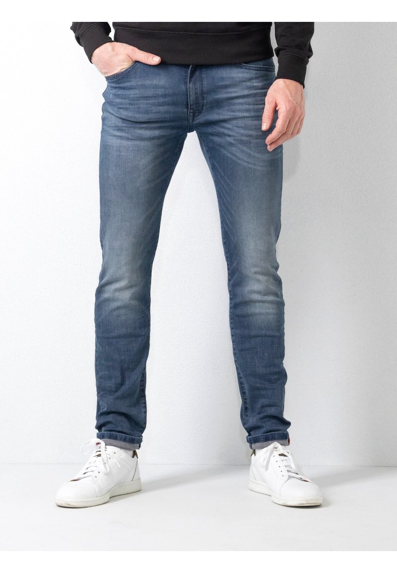 Herrarnas smalpassande jeans i medelblå denim med tvättad yta, försedda med framfickor och slitna detaljer, tillsammans med vita sneakers.