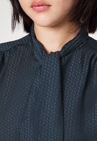 Tummansininen pusero, jossa hienovarainen geometrinen kuvio, solmittava kaula-aukko ja pehmeä, teksturoitu kangas. Lähikuva, joka korostaa yksityiskohtia.