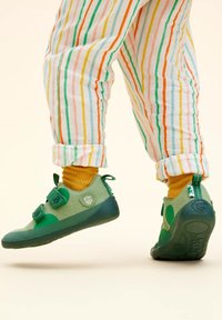 Groene stoffen schoenen met twee klittenbanden, ronde neuzen en gestructureerde zolen. Draagt het met gele sokken en gestreepte broek in meerdere kleuren.