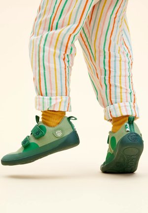 Grüne Stoffschuhe mit zwei Klettverschlüssen, runden Zehen und strukturierten Sohlen. Kombiniert mit gelben Socken und gestreiften Hosen in mehreren Farben.