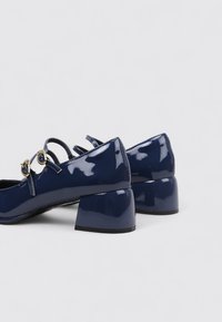 Zapatos de charol azul marino con punta redonda y tacón cuadrado, con detalles de correas y acentos dorados. Acabado suave y brillante.