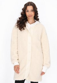 Cappotto teddy color crema, morbido e fluffy, con chiusura a bottoni e cappuccio. Caratterizzato da una texture soffice, maniche lunghe e fodera interna a contrasto.