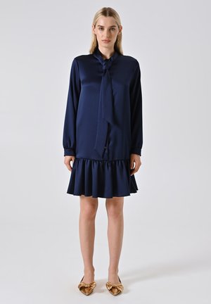 Femme avec des cheveux blonds raides portant une robe longue à manches longues bleu marine avec un nœud à col et un ourlet à volants, associée à des ballerines dorées pointues avec des nœuds.