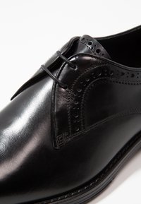 Chaussure habillée en cuir noir avec une finition lisse, présentant des perforations décoratives le long du bord, un design à lacets et un bout arrondi.