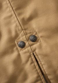 Tkanina v barvi rjave kože z gladko teksturo vsebuje navpični šiv, dve kovinski sponki za pritrditev in kontrastne detajle šivanja.