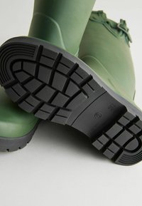 Groene rubberen regenlaarzen met een getextureerde zwarte rubberen zool, voorzien van diepe noppen voor grip. Het ontwerp heeft een ronde teen en een hoge schacht.