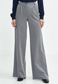 Szare spodnie o szerokich nogawkach wykonane z teksturowanej tkaniny, z przednimi zakładkami, zapięciem na guzik i wysokim stanem, zestawione z czarnymi butami.