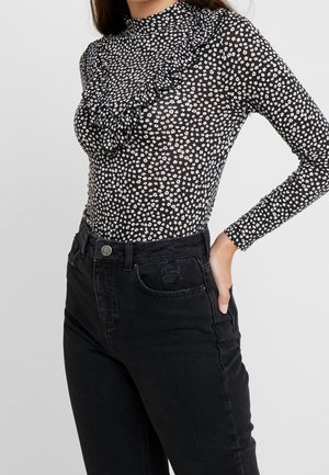 Vrouw draagt een zwarte top met lange mouwen, kleine witte bloemen en ruches, gecombineerd met zwarte high-waist jeans, hand in de zak.