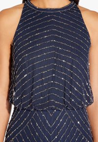 Haut sans manches bleu marine avec un design évasé, orné de motifs en zigzag de sequins dorés et d'un col haut. Texture de tissu douce et fluide.