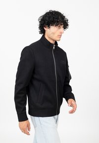 Zwarte bomberjack met hoge kraag, ritssluiting aan de voorkant, zijzakken, textuurstof en een klein reliëflogo op de borst.