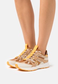 Beige och tan atletiska skor med gula accenter, mesh- och mockamaterial, intrikat snörningssystem och strukturerad sula.