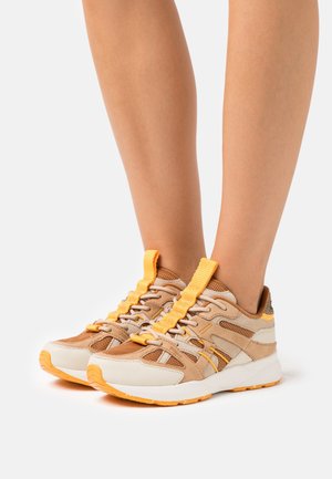 Scarpe da ginnastica beige e tan con accenti gialli, materiali in rete e suede, sistema di allacciatura complesso e design della suola testurizzato.
