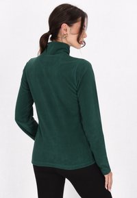 Pullover en polaire vert foncé à manches longues avec un col haut et une texture lisse. Il présente un design ajusté et des détails de couture subtils.